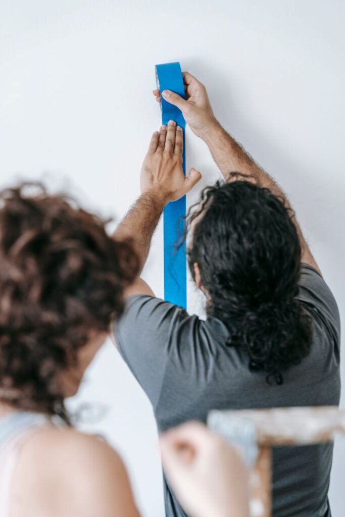 Two adults applying blue adhesive tape during a home renovation indoors.