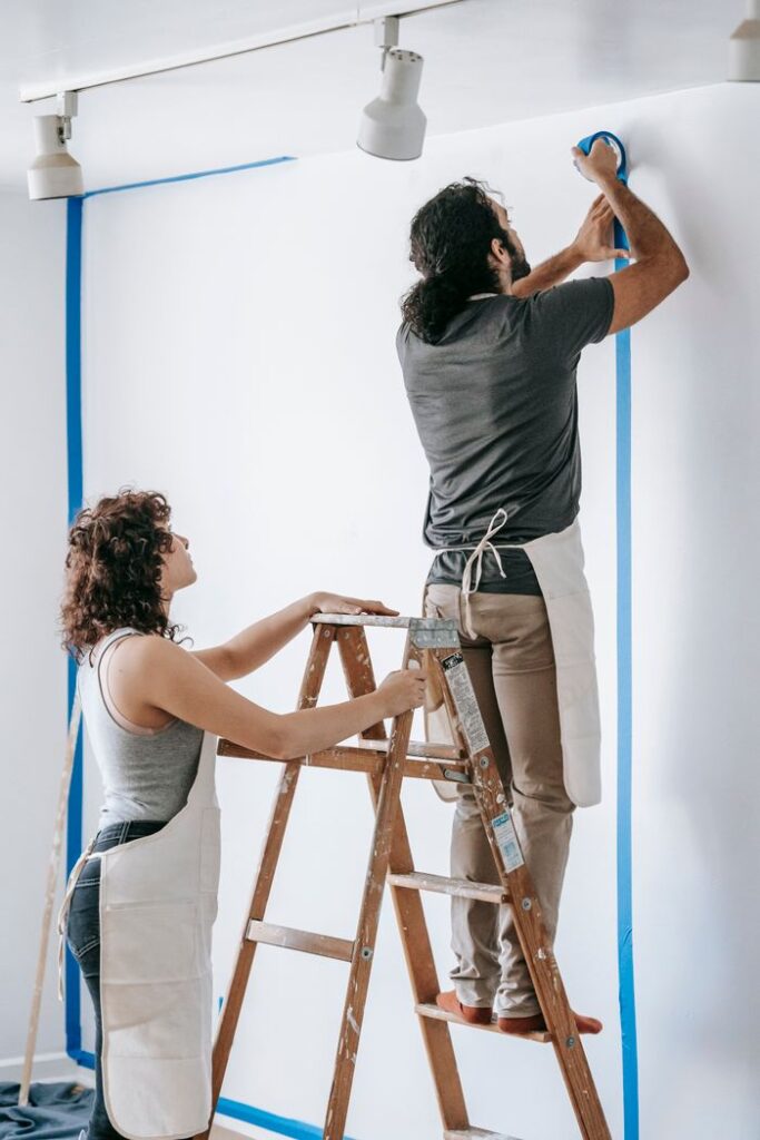 Couple working together on home renovation, taping wall with stepladder.